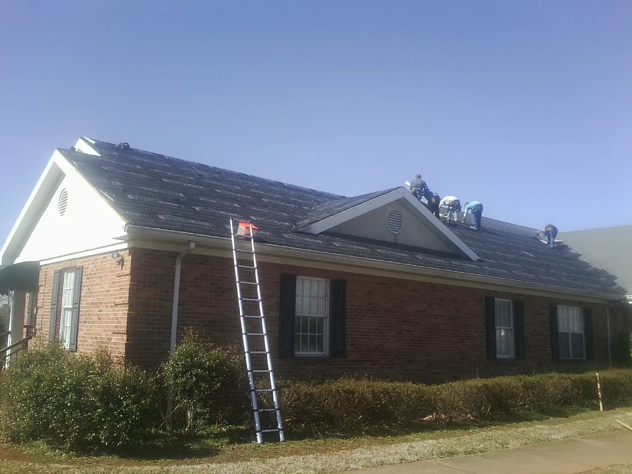 Crew tearing shingles off a brick ranch roof with a ladder set — Metal Roof Repair work by our crew in Union Springs